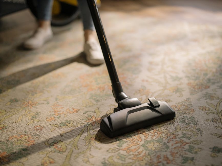 A woman vacuuming a patterned area rug in a living room with wooden flooring and a brown leather sofa. She is holding a yellow canister vacuum cleaner, cleaning the light-colored, floral-design carpet