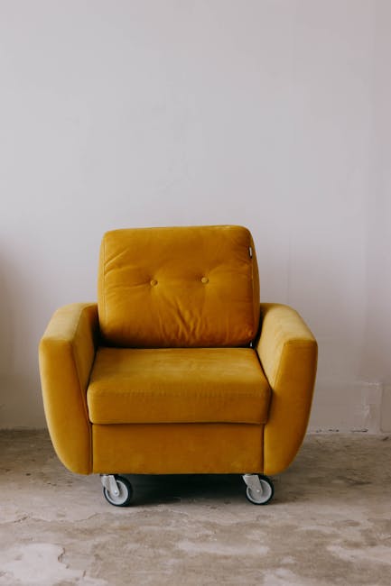 A person wearing a black leather jacket is using a black handheld vacuum cleaner to deep clean a yellow and white patterned cushion placed on a dark fabric sofa in a well-lit indoor setting. The scene